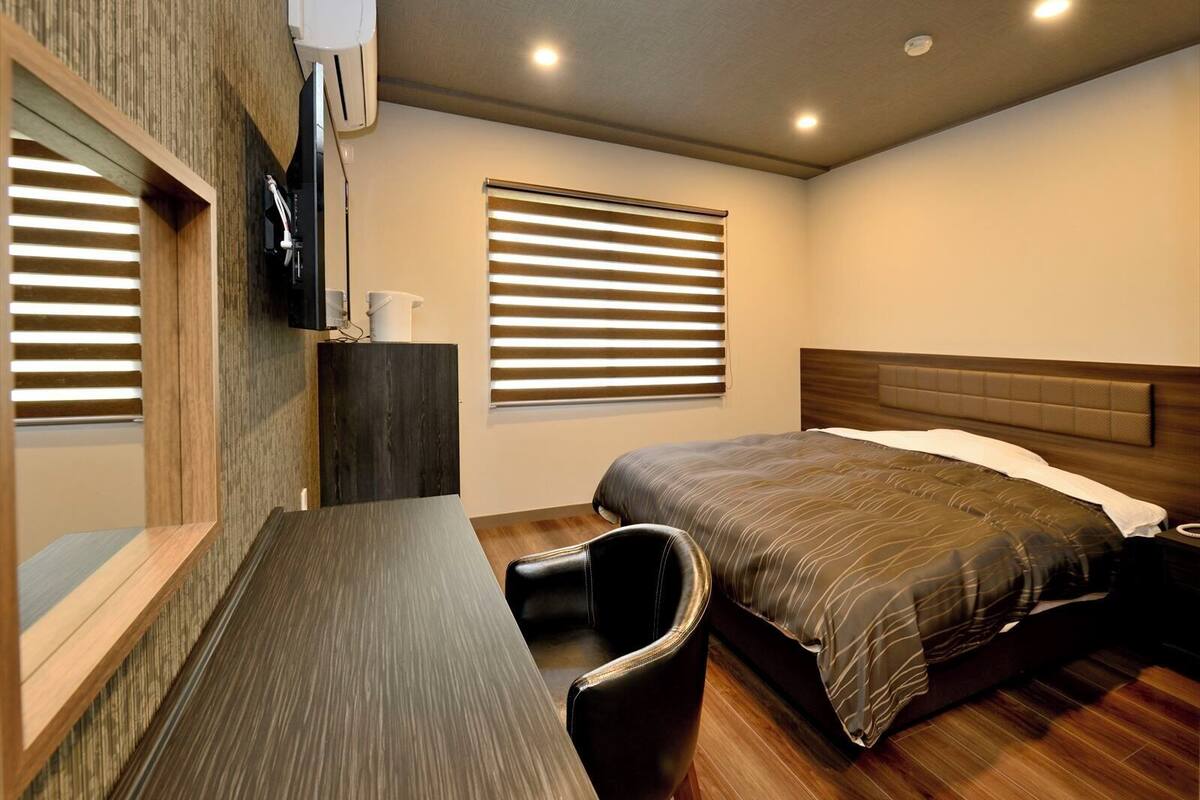 Een slaapkamer met een bed, bureau, stoel, televisie en een raam met zonwering.