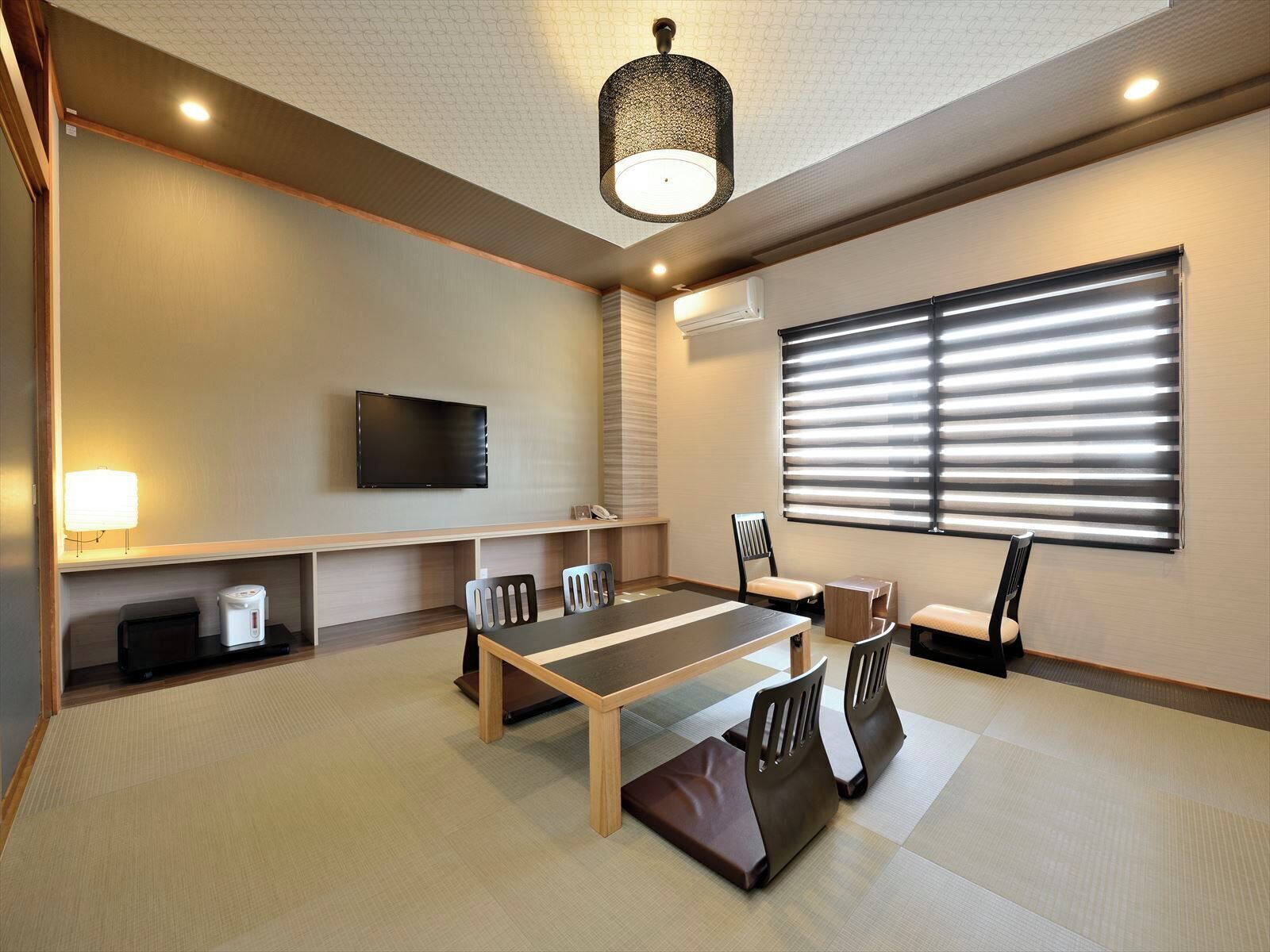A modern meeting room with a flat-screen TV, a long table, chairs, a lamp, and a large window with blinds.