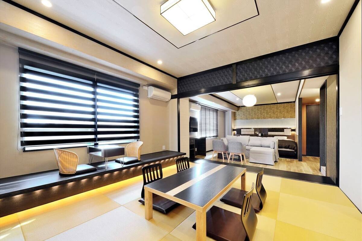 Een moderne woonkamer met een eethoek, grote ramen met zonwering, een bank en een televisie.
