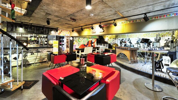 Un restaurant avec des sièges rouges et noirs, une scène avec des instruments de musique et un tableau affichant le menu.