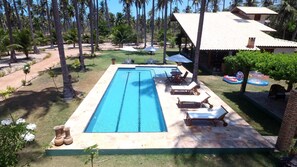 A rectangular swimming pool with a tiled deck, surrounded by palm trees and a building with a tiled roof.