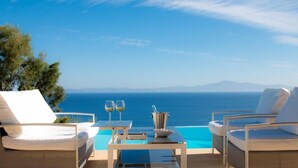 A poolside area with lounge chairs, a table with two glasses of wine, and a view of the sea and mountains.
