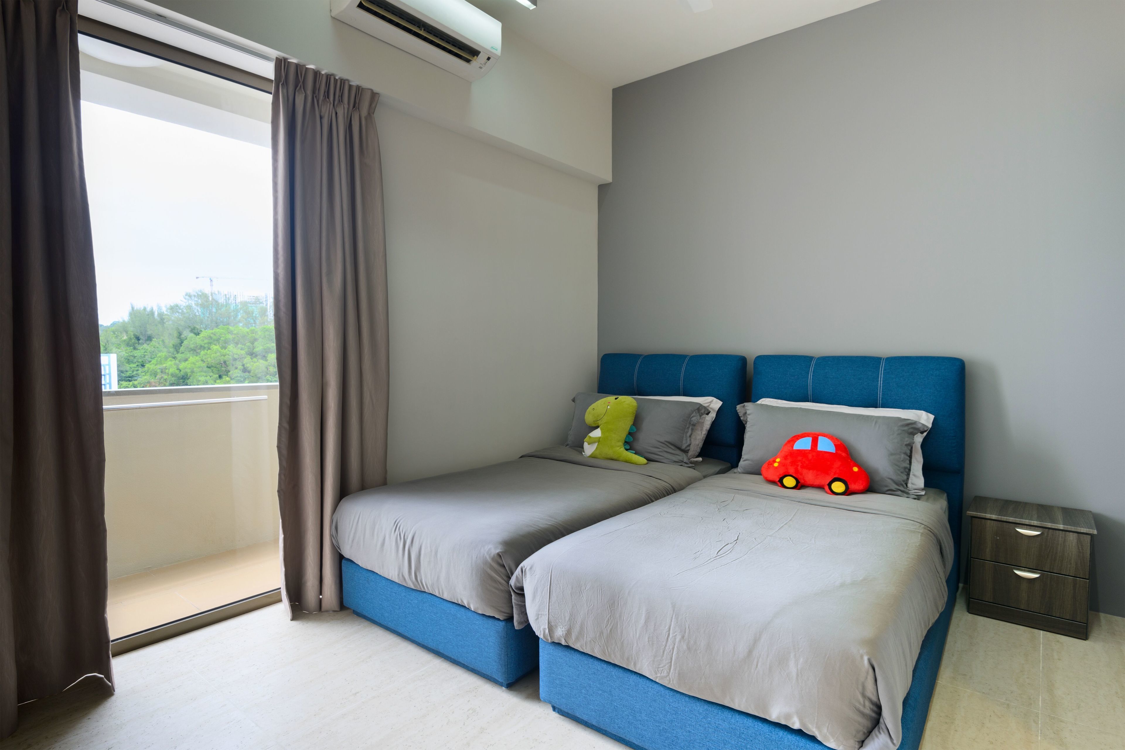 A bedroom with two beds, a window with curtains, a nightstand, and a view of greenery outside.