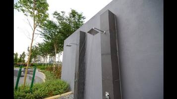 A modern outdoor shower with a vertical water feature against a grey wall, surrounded by greenery.