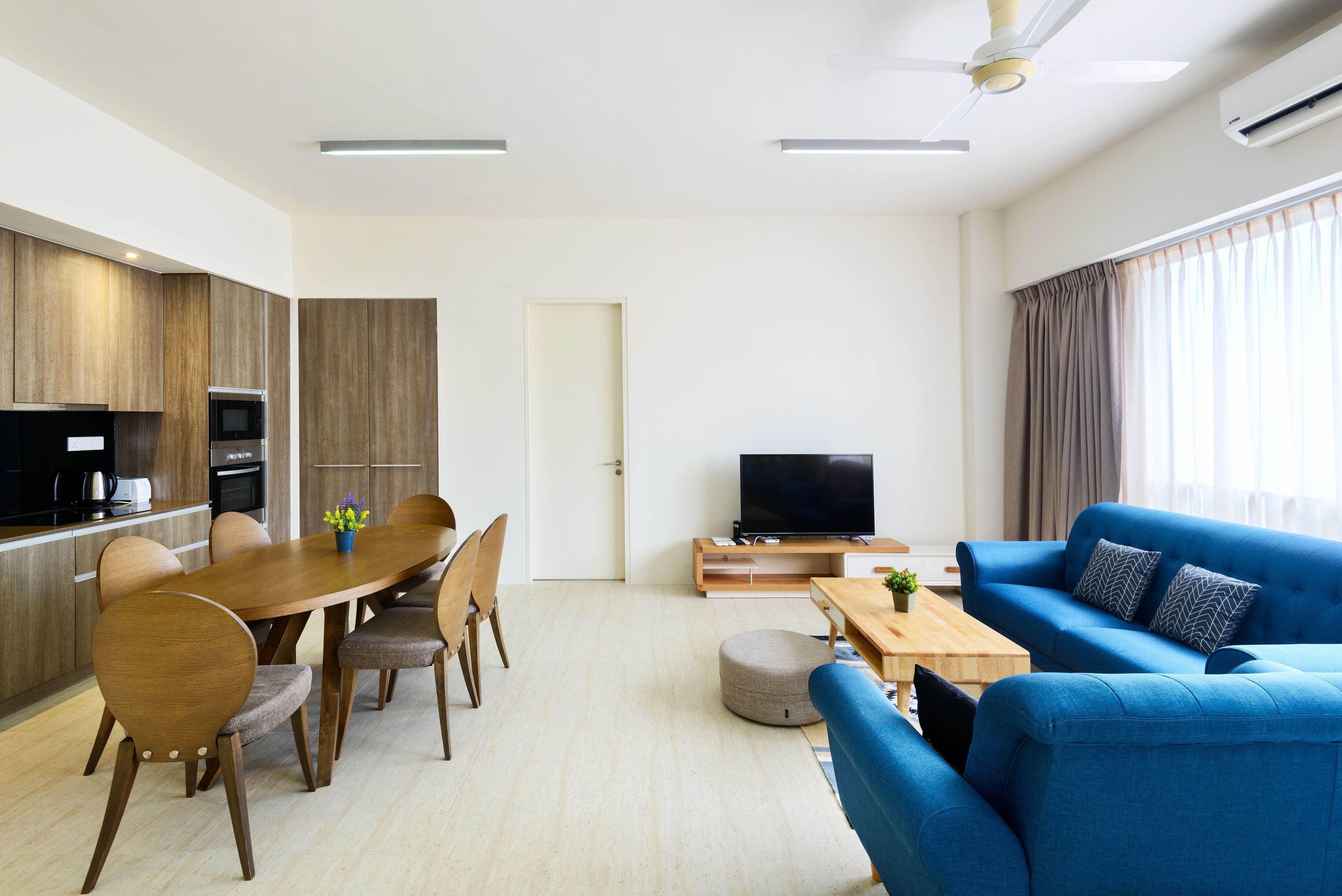 Een moderne woonkamer met een blauwe bank, een houten eettafel en stoelen. Er is een televisie op een houten kast en een keuken met kasten en apparatuur.