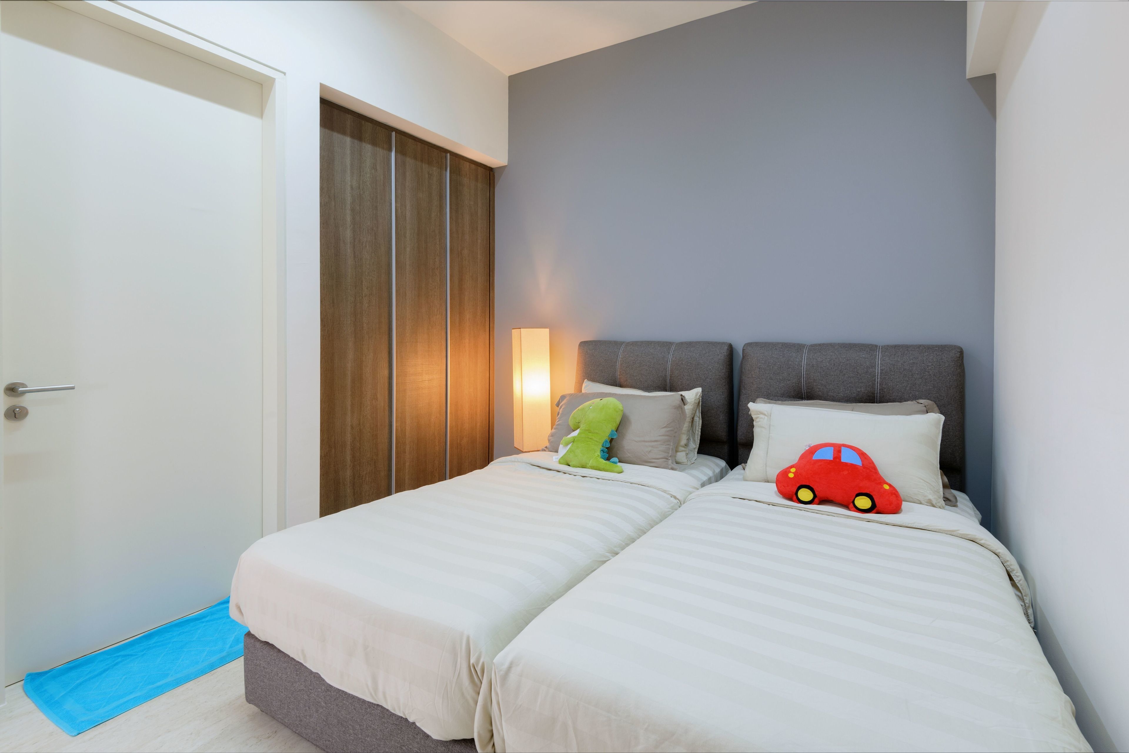 Una camera da letto con due letti, un armadio in legno, un tappetino blu all'ingresso e una lampada fissata alla parete.