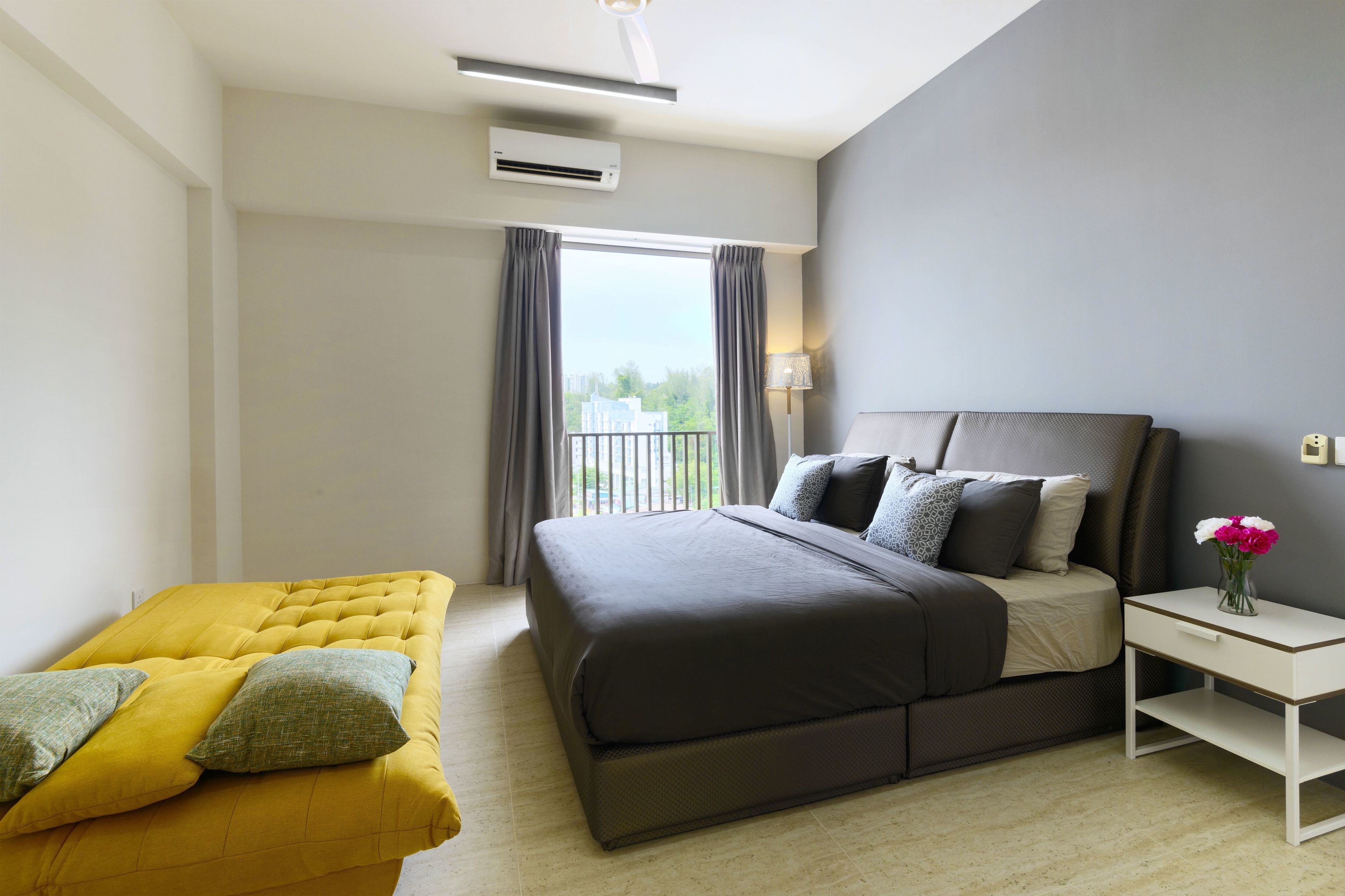 A modern hotel room with a large bed, a yellow sofa, a bedside table with a vase of flowers, and a view of the outdoors through a window.