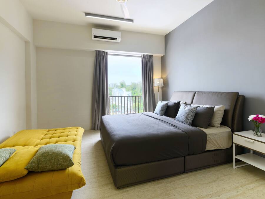 A modern hotel room with a large bed, a yellow sofa, a bedside table with a vase of flowers, and a view of the outdoors through a window.