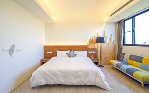 A modern hotel room with a large bed, a wooden headboard, a bedside table with a lamp, and a colorful sofa.