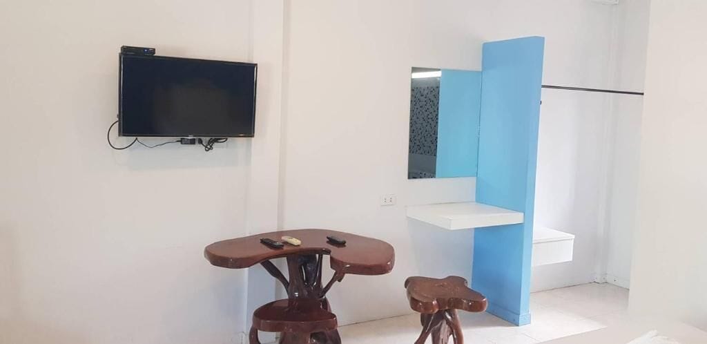Quarto com televisão de tela plana na parede, uma pequena mesa de madeira com dois banquinhos e uma parede azul com um estante branco.