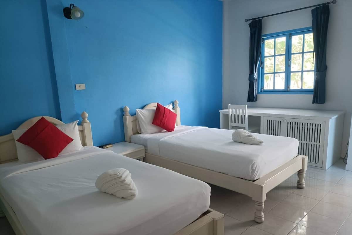 Quarto de hotel com duas camas, cada uma com roupa de cama branca e almofadas vermelhas, uma mesa pequena, uma cadeira e uma janela com cortinas.