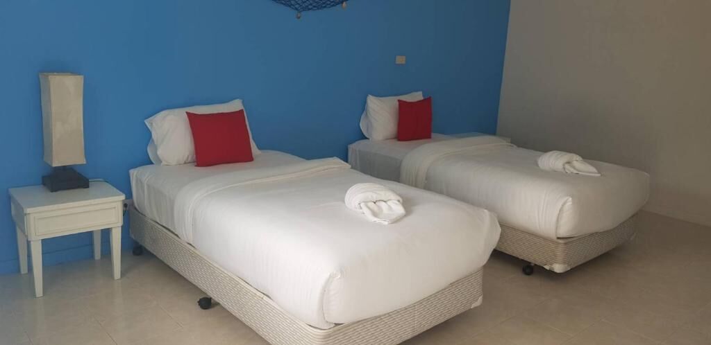 Een hotelkamer met twee bedden, elk voorzien van witte lakens en rode kussens. Er is een klein nachtkastje met een lamp en een wandplank boven de bedden.