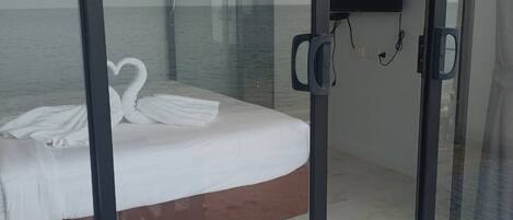 A bedroom with a bed, a TV, and a view of the sea through a glass door.