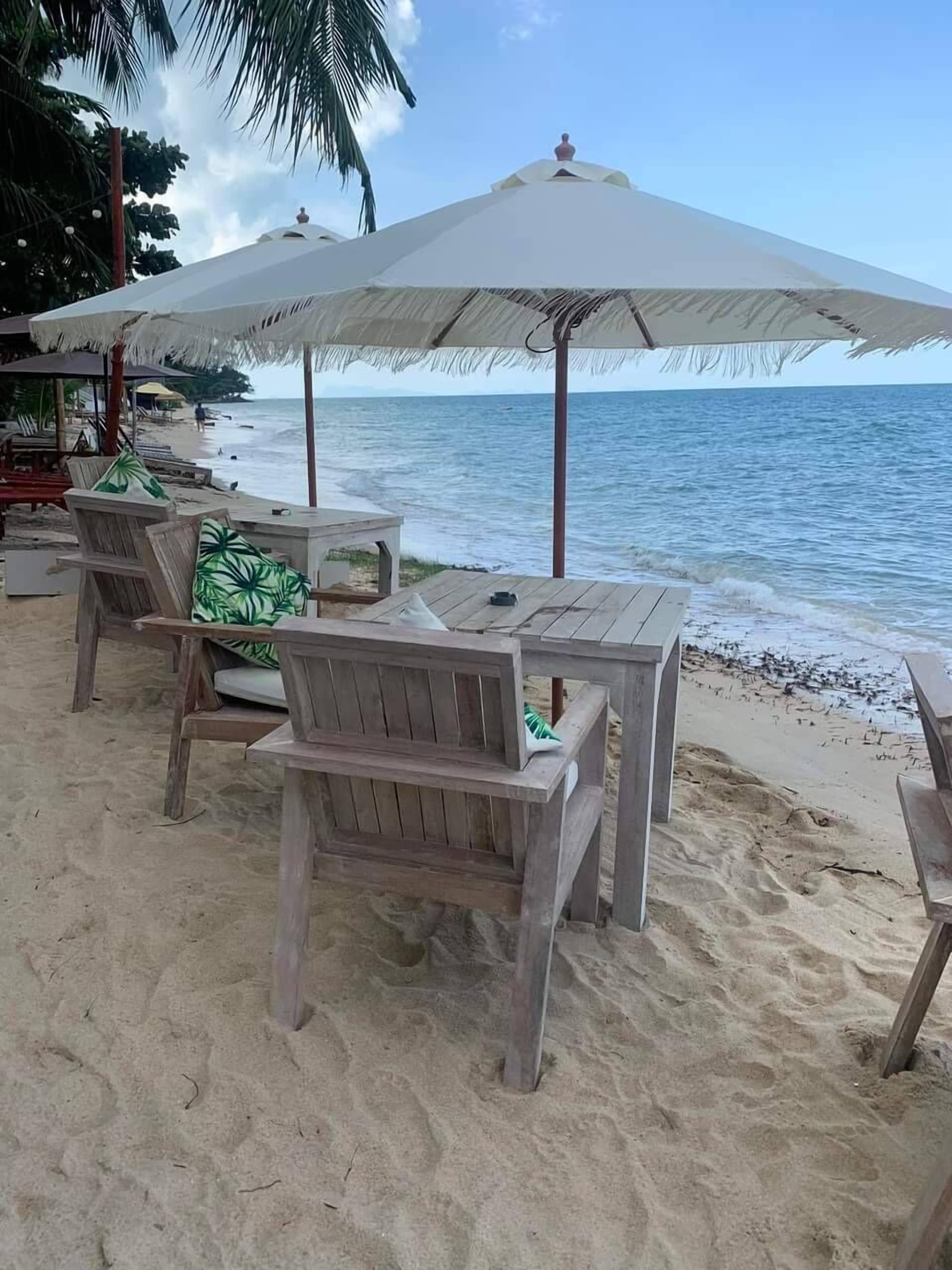 A beachside seating area with wooden chairs and a table, an umbrella, and a view of the ocean.