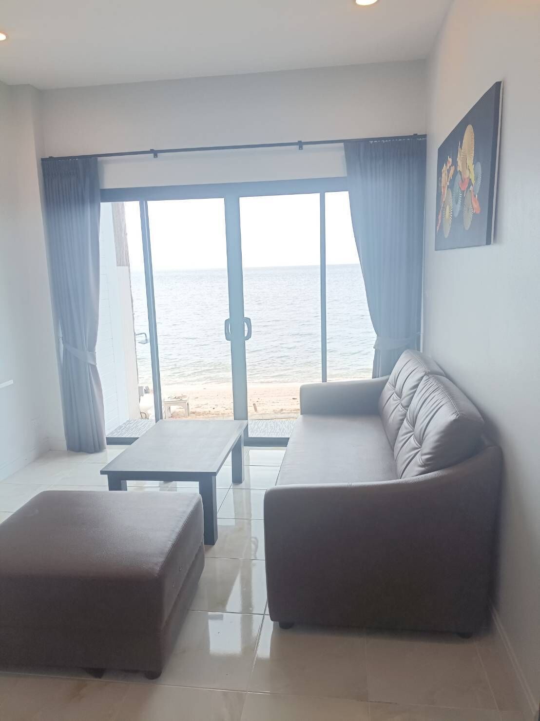 Un salon moderne avec un canapé, un pouf et une table, donnant sur une plage et offrant une vue dégagée sur la mer.