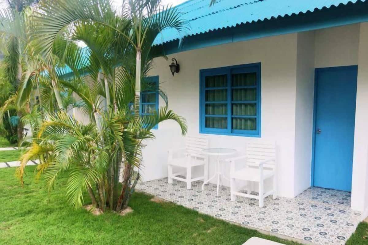 Um edifício branco com porta e janelas azuis, um pequeno pátio com cadeiras brancas e uma palmeira.