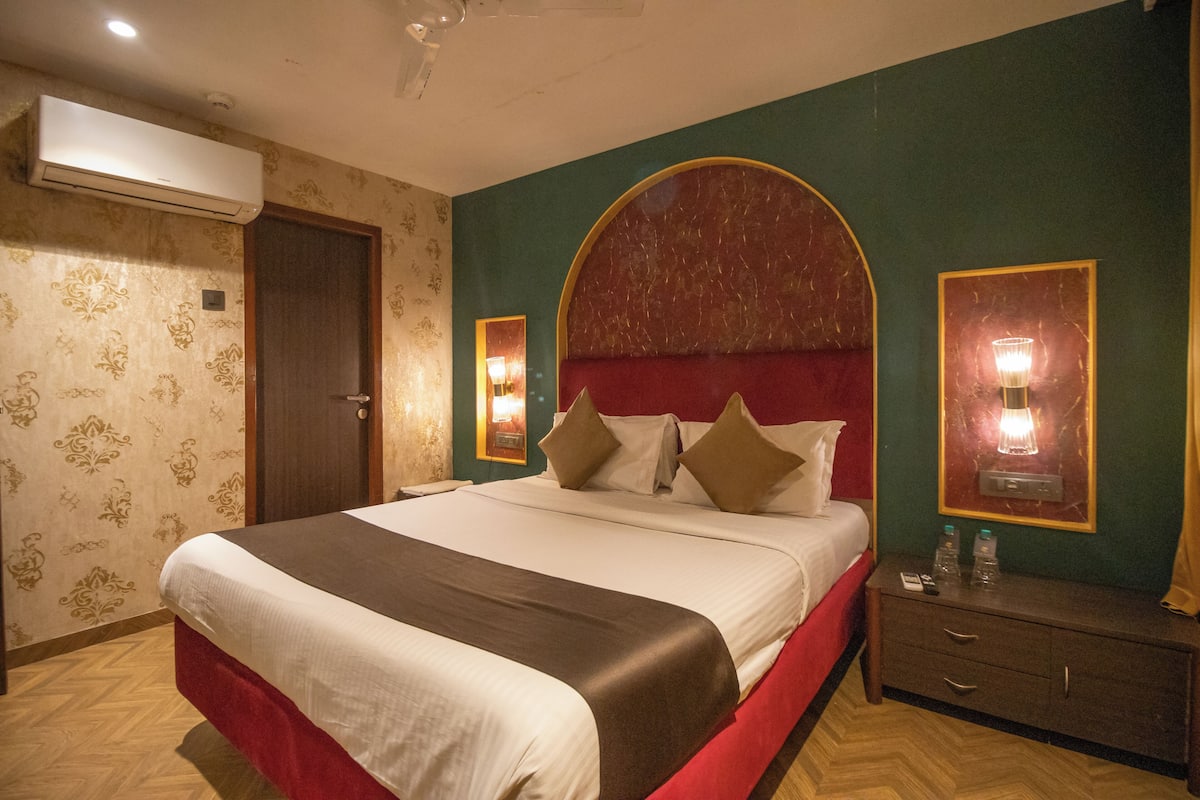 A hotel room with a large bed, a wooden floor, an air conditioning unit, a door, a nightstand with a lamp, and a mirror.