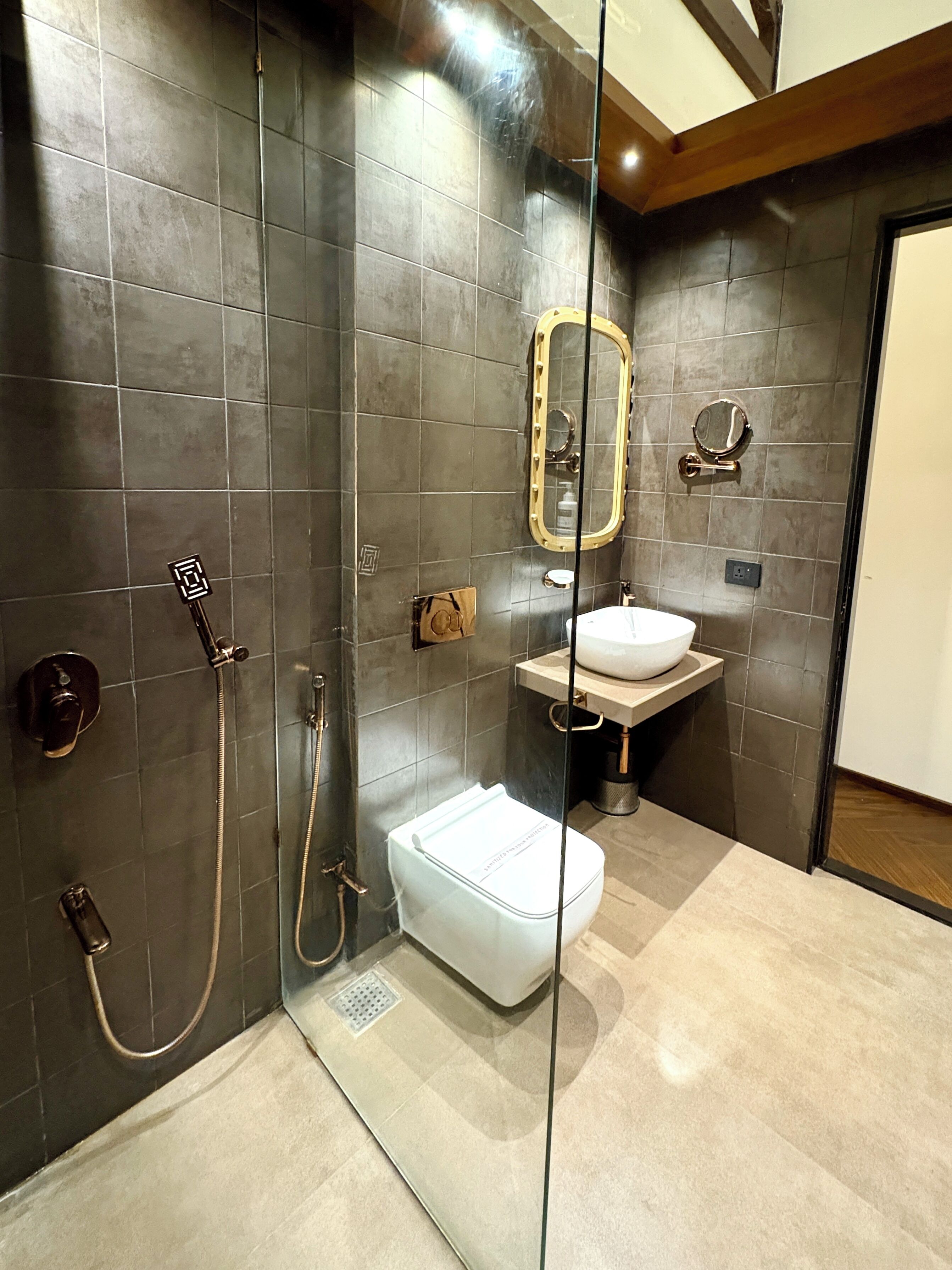 Un bagno moderno con pareti piastrellate di colore scuro, box doccia in vetro, un water bianco, un lavandino con specchio dorato e un soffitto in legno.