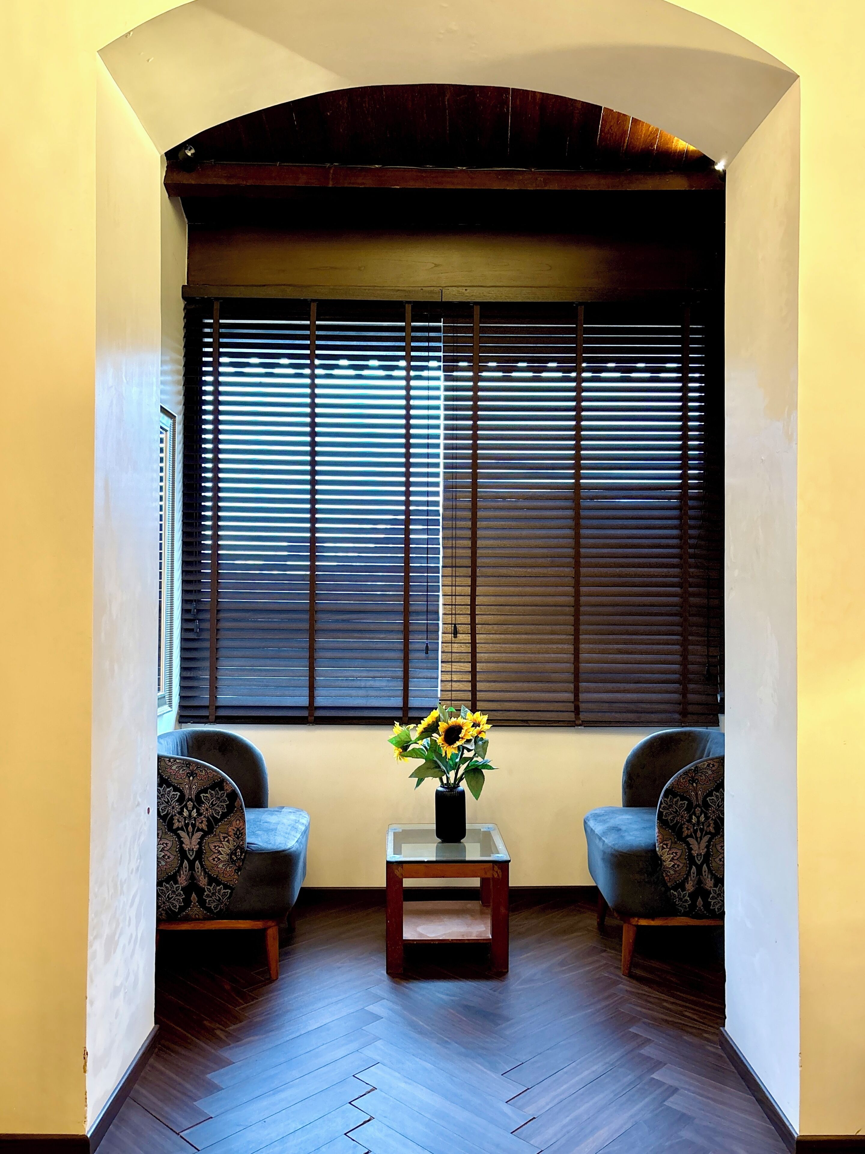 A room with a wooden floor, two patterned armchairs, a small table with a vase of flowers, and a large window with blinds.