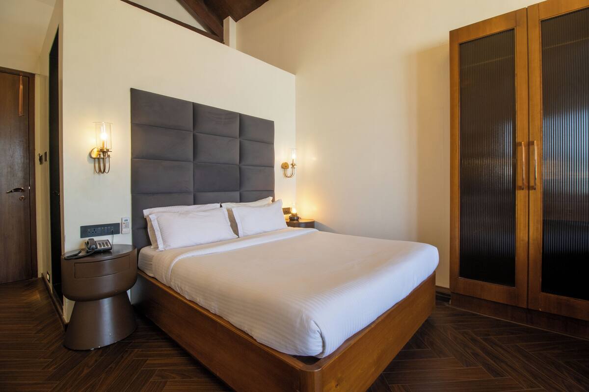 A modern bedroom with a large bed, bedside lamps, a wooden wardrobe, and a nightstand with a phone and charger.