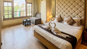 A spacious bedroom with a large bed, a seating area, and a view of a cityscape through arched windows. - Gulf Hotel Colaba (Mumbai)