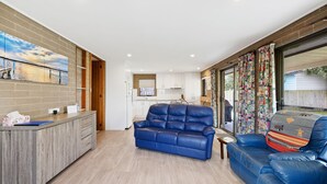 Uma sala de estar moderna com um sofá de couro azul, uma mesa de centro de madeira e uma área de cozinha com armários brancos e revestimento de tijolos.