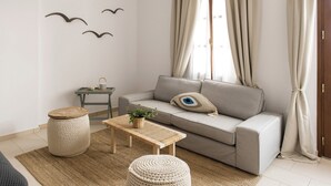 A modern living room with a grey sofa, a round wooden coffee table, a woven ottoman, and a small side table with a plant. - Syros Wellness Luxury Suites (Syros)