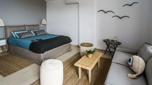A modern bedroom with a bed, a grey sofa, a wooden coffee table, and a white pouf. - Syros Wellness Luxury Suites (Syros)