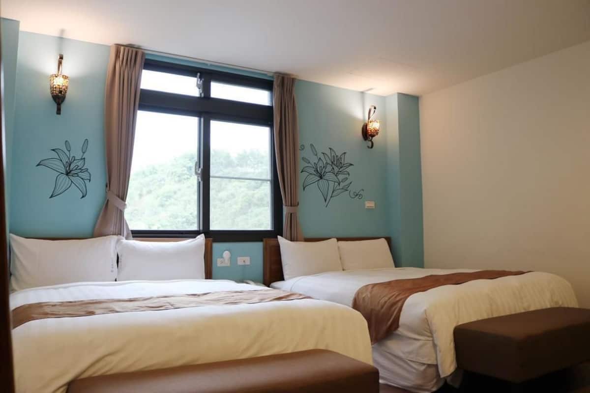 A hotel room with two beds, a window with curtains, wall-mounted lights, and a view of greenery outside.