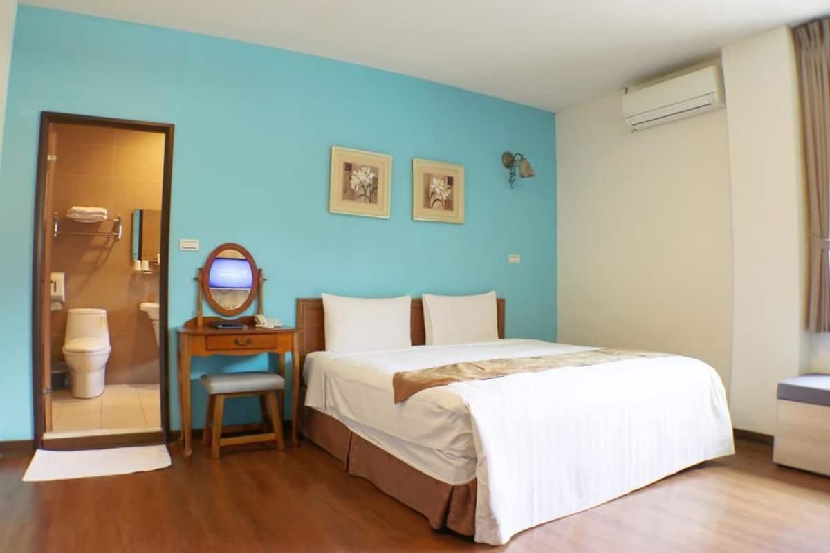 A hotel room with a bed, a desk with a chair, a mirror, and two framed pictures on the wall.