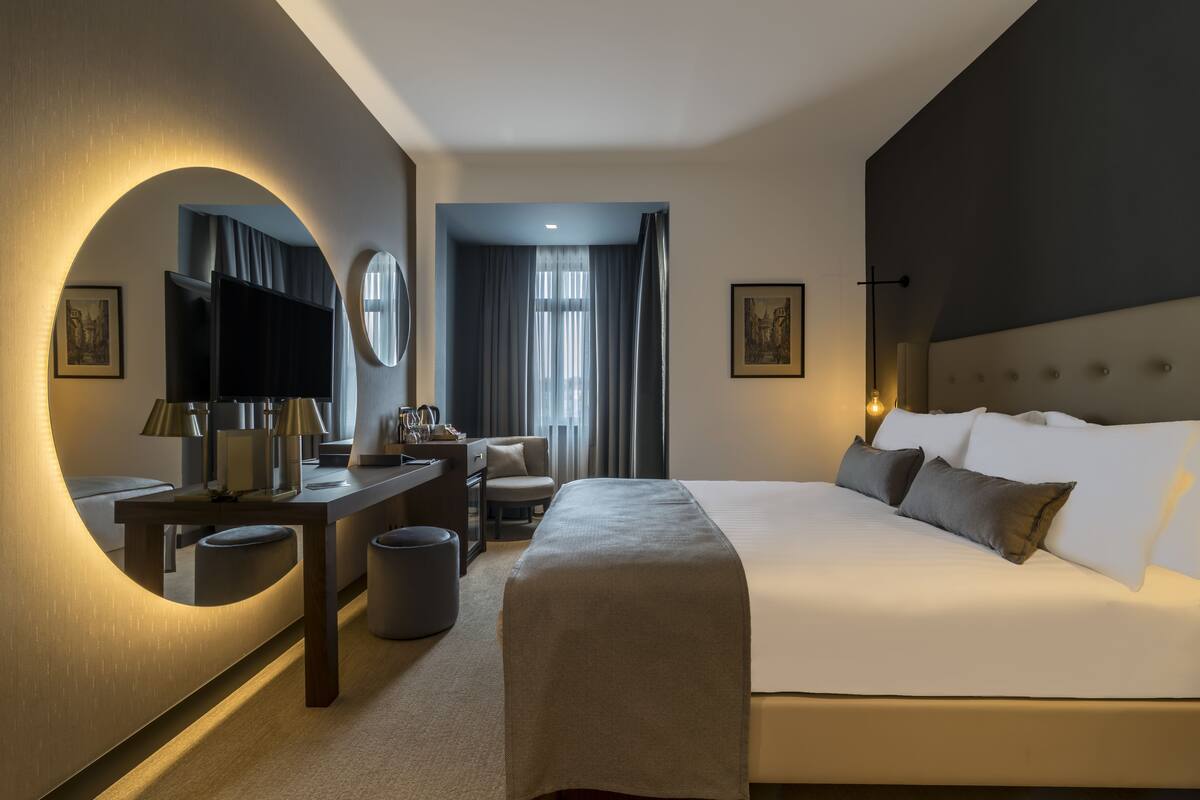 A modern hotel room with a large bed, a desk with a flat-screen TV, a comfortable armchair, and a large mirror.