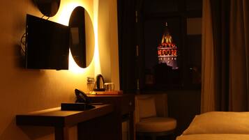 A hotel room with a bed, a desk with a lamp, a chair, a TV mounted on the wall, and a view of a building through the window.