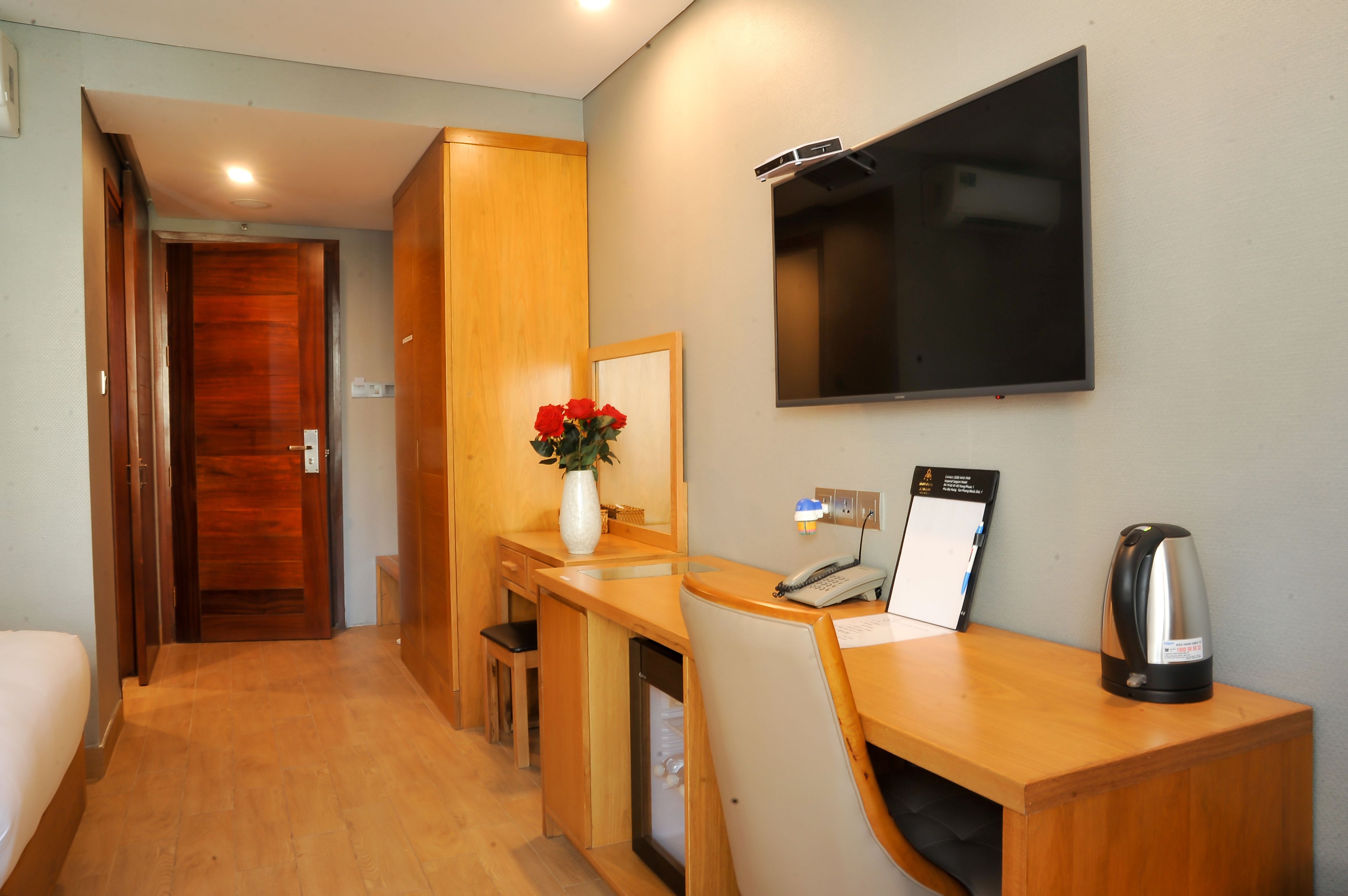 A hotel room with a wooden desk, a chair, a small table, a vase with red flowers, a kettle, and a flat-screen TV mounted on the wall.