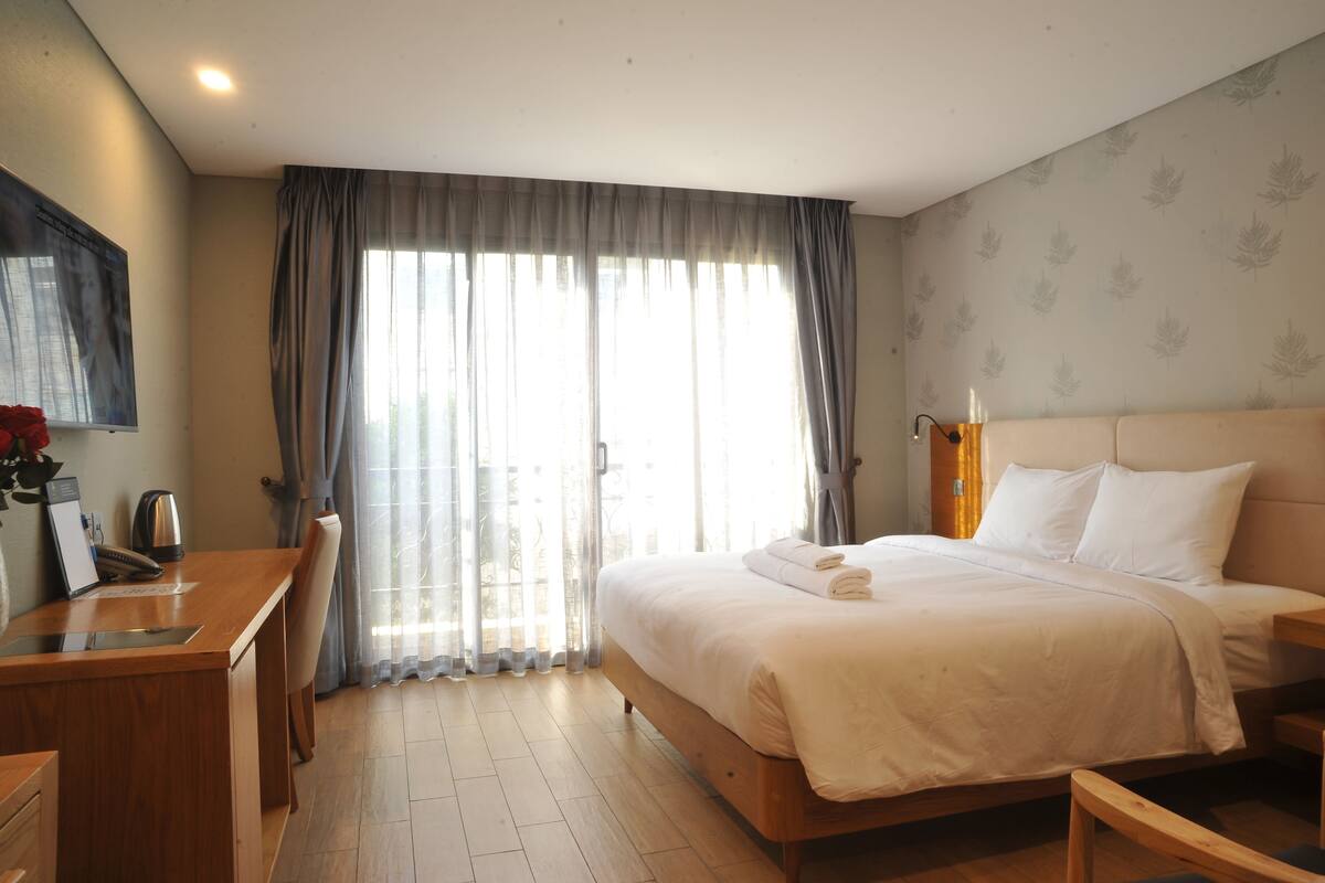 A hotel room with a bed, a desk, a chair, a coffee maker, and a view of the outside through large windows.