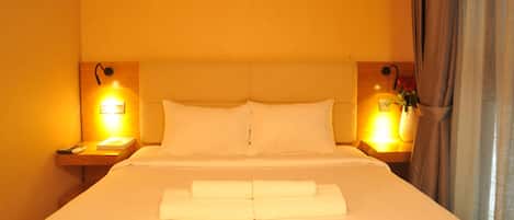 A hotel room with a large bed, two bedside tables, a nightstand, a wall-mounted lamp, and a window with curtains.