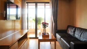 A living room with a wooden TV stand, a black leather sofa, a wooden coffee table with a vase of flowers, and a sliding glass door leading to an outdoor balcony. - Imperial Saigon Hotel (Ho Chi Minh City)