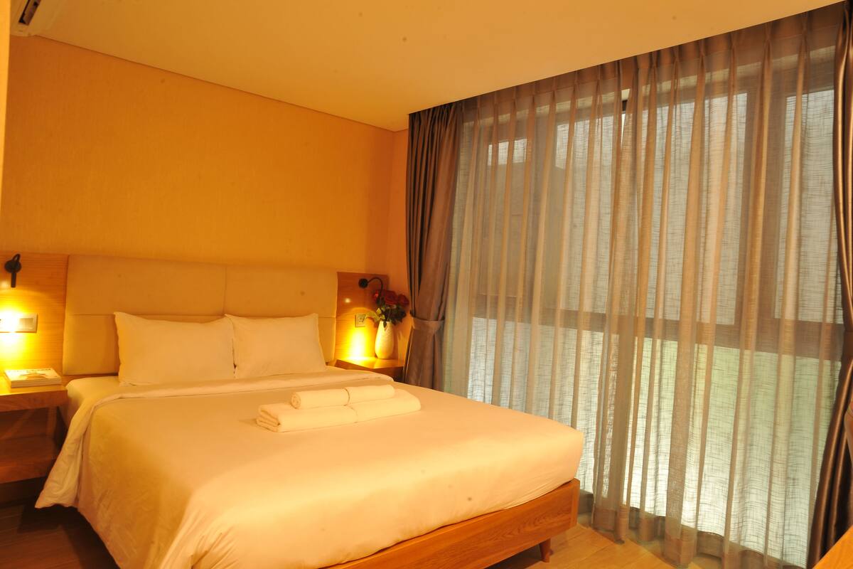 A hotel room with a large bed, two bedside lamps, a nightstand, and a window with sheer curtains.