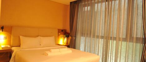 A hotel room with a large bed, two bedside lamps, a nightstand, and a window with sheer curtains.