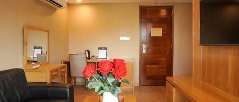 A modern hotel room with a wooden floor, a black leather sofa, a wooden coffee table with a white vase of red roses, a wooden desk with a computer, and a flat-screen TV mounted on the wall.