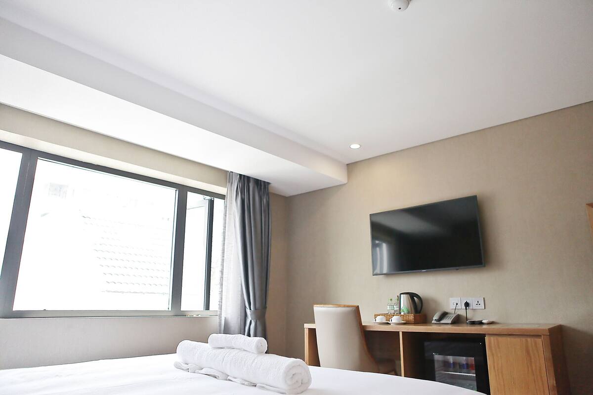 A hotel room with a bed, a desk with a flat-screen TV, a chair, and a window with curtains.