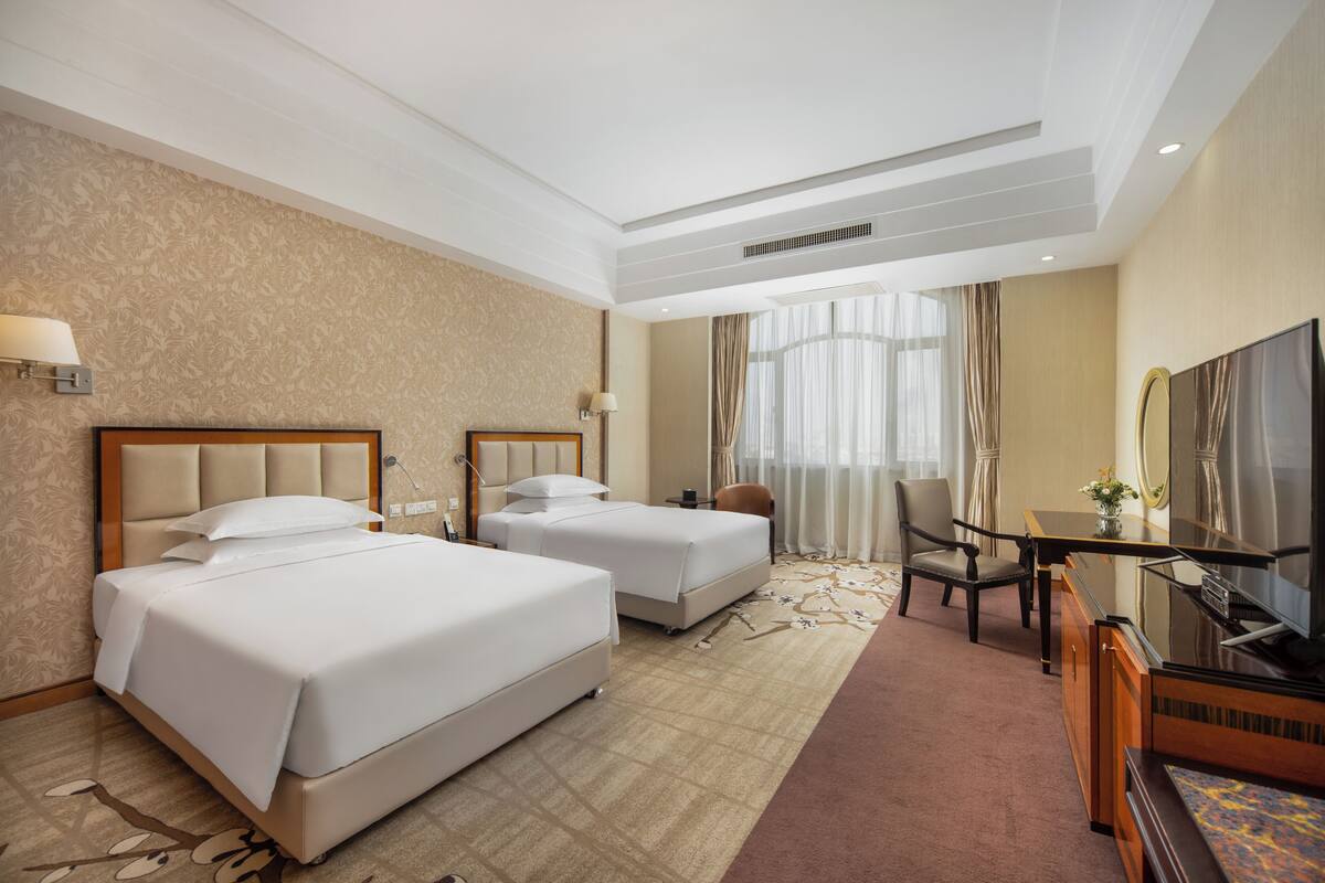 Habitación de hotel con dos camas, un escritorio, una silla, un televisor y un ventanal grande con cortinas.
