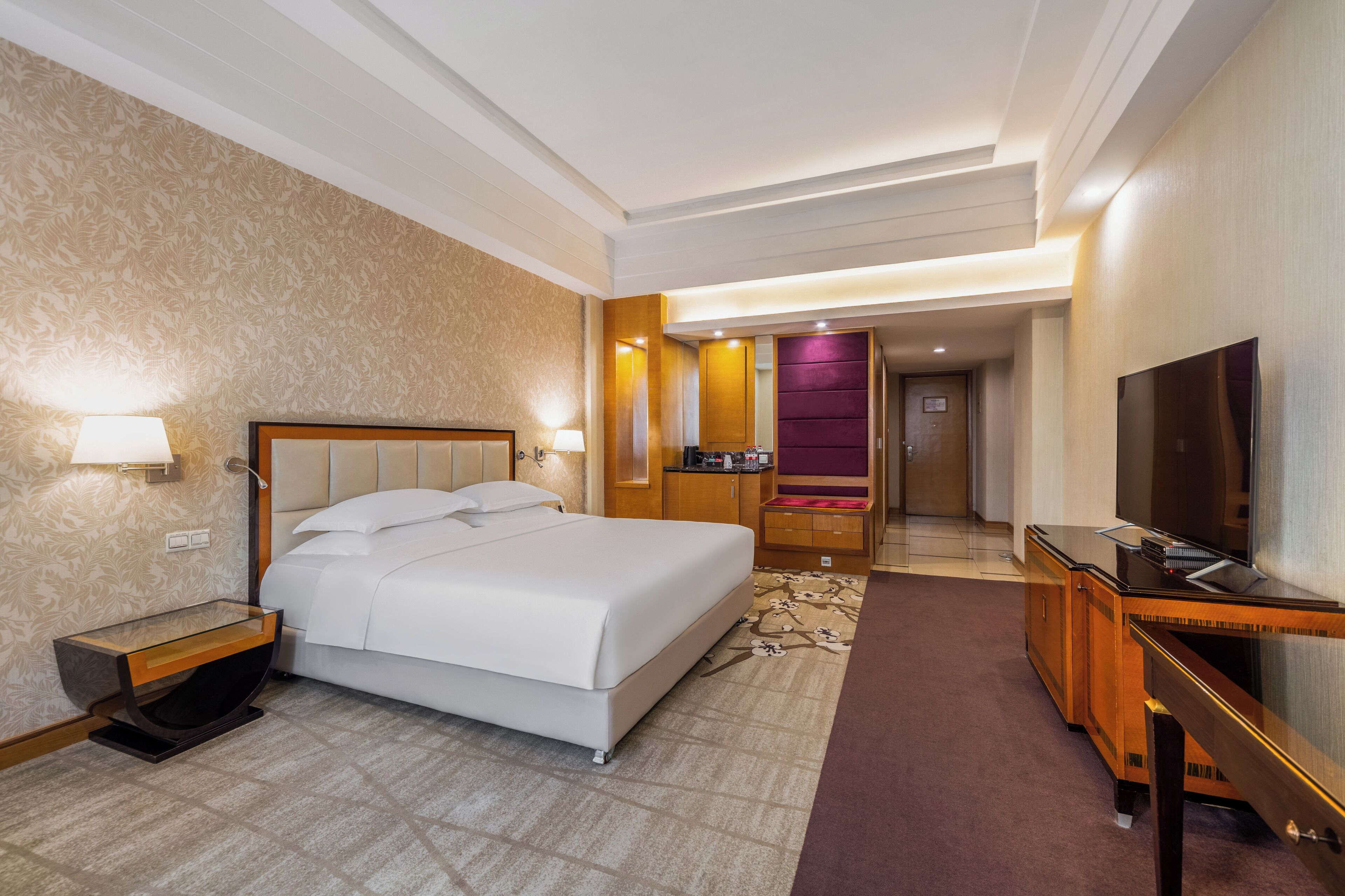 A hotel room with a bed, bedside table, lamp, TV, and a desk with a chair.