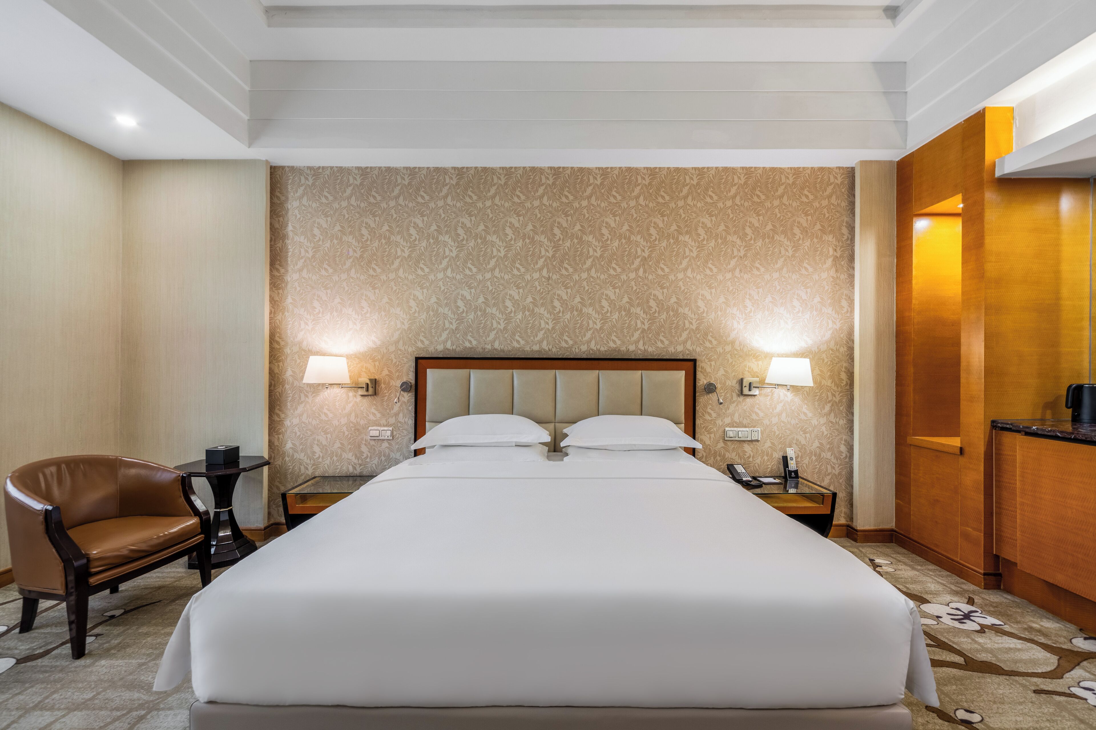 A hotel room with a large bed, two bedside tables with lamps, a leather armchair, and a wooden desk with a coffee maker.
