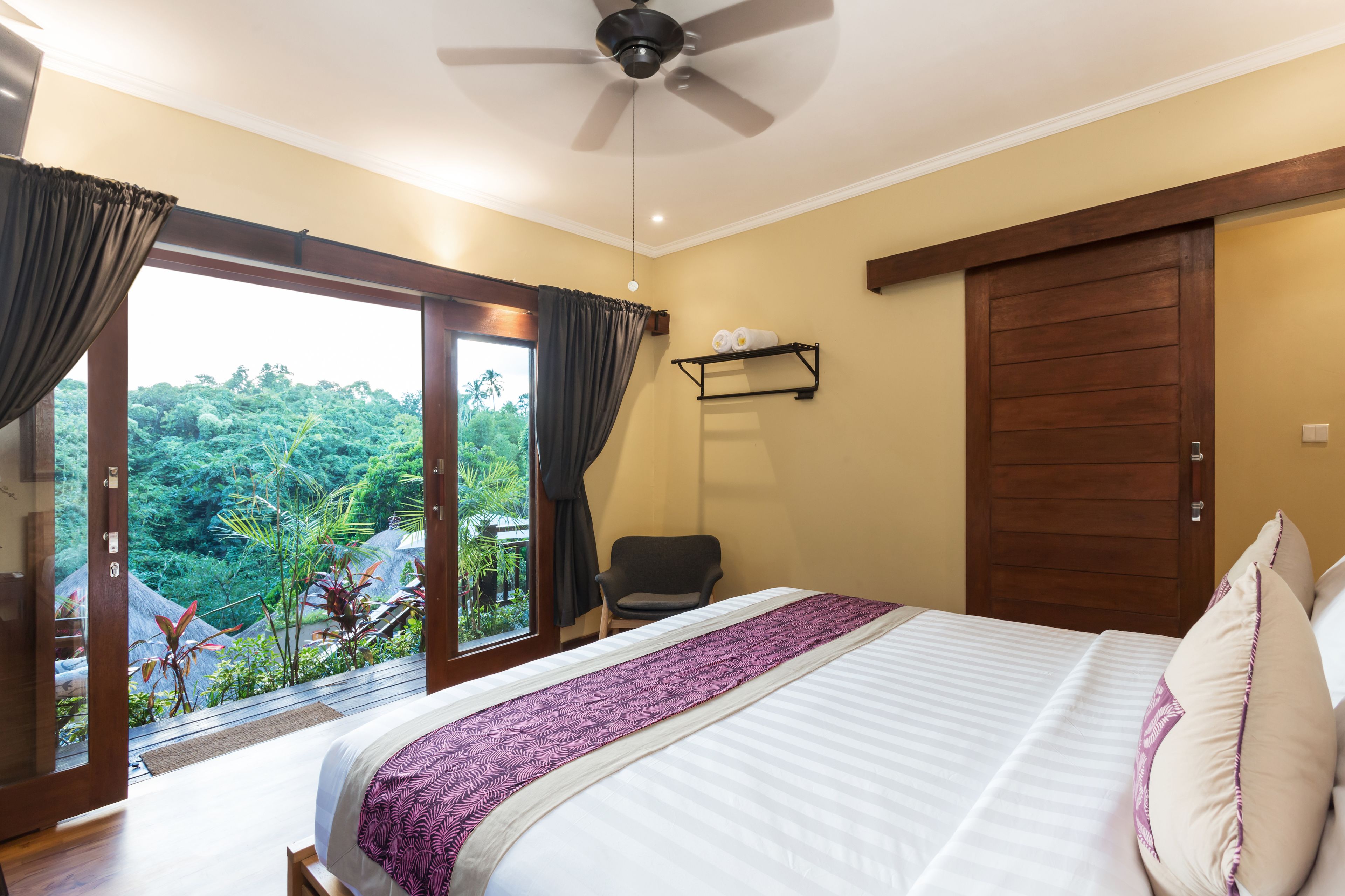 Un dormitorio con cama, ventilador de techo, ventana con cortinas, una silla y una puerta.