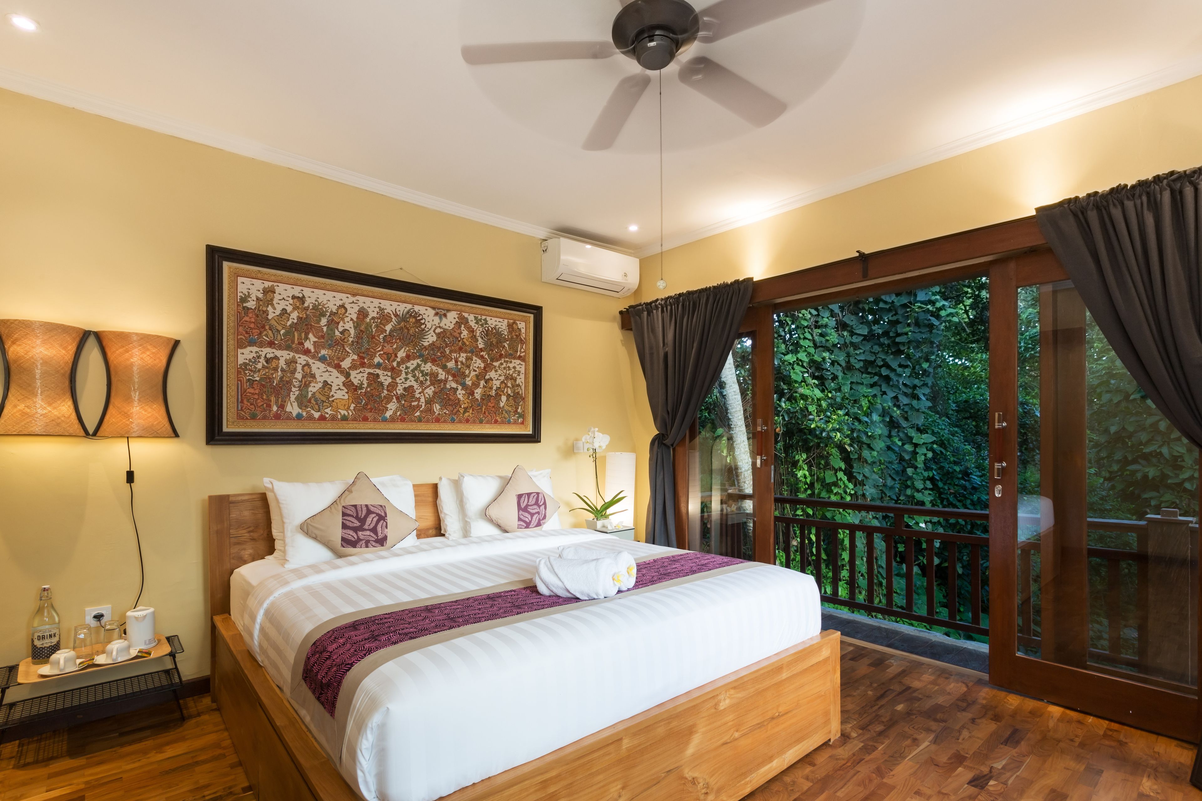 A well-appointed bedroom with a large bed, a ceiling fan, a wall-mounted air conditioner, and a view of lush greenery through a sliding glass door.