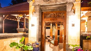 A traditional building entrance with a wooden door and stone carvings, surrounded by potted plants and a small wooden stool. - The Pari Sudha (Ubud)