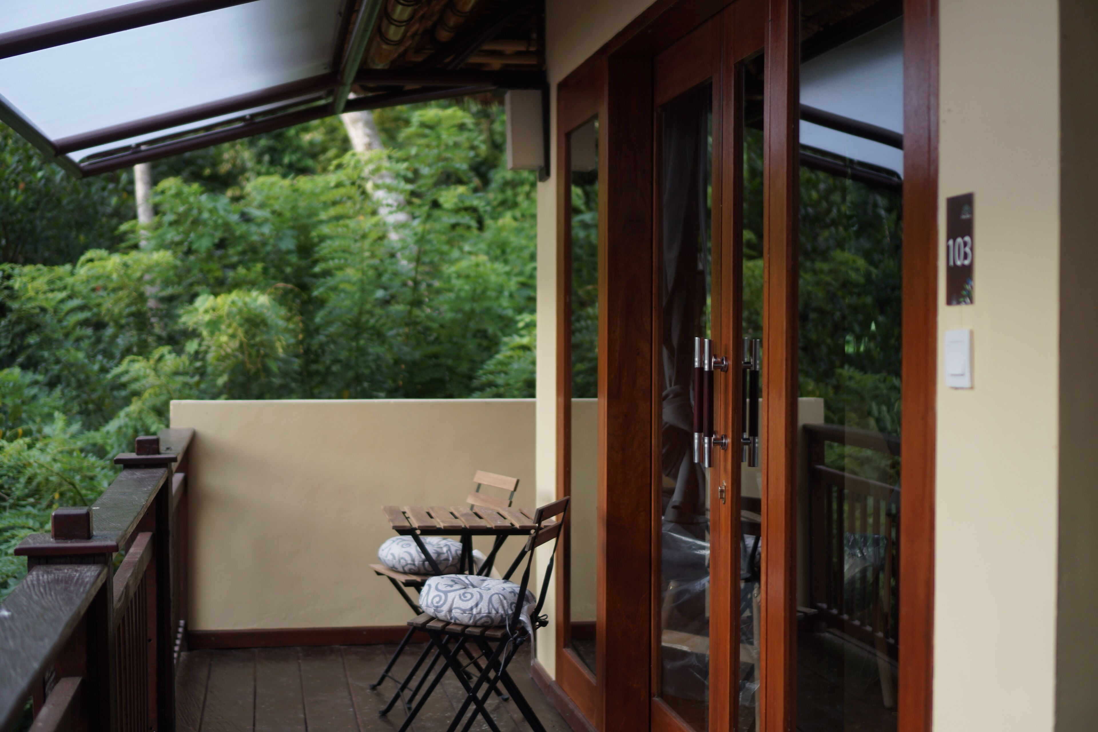A balcony with a wooden table and two chairs, a glass door, and a view of lush greenery.