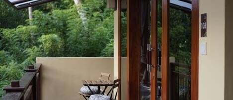 A balcony with a wooden table and two chairs, a glass door, and a view of lush greenery.