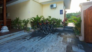 A building entrance with a stone-paved path leading to a wooden door, surrounded by potted plants and bicycles parked against the wall. - The Pari Sudha (Ubud)