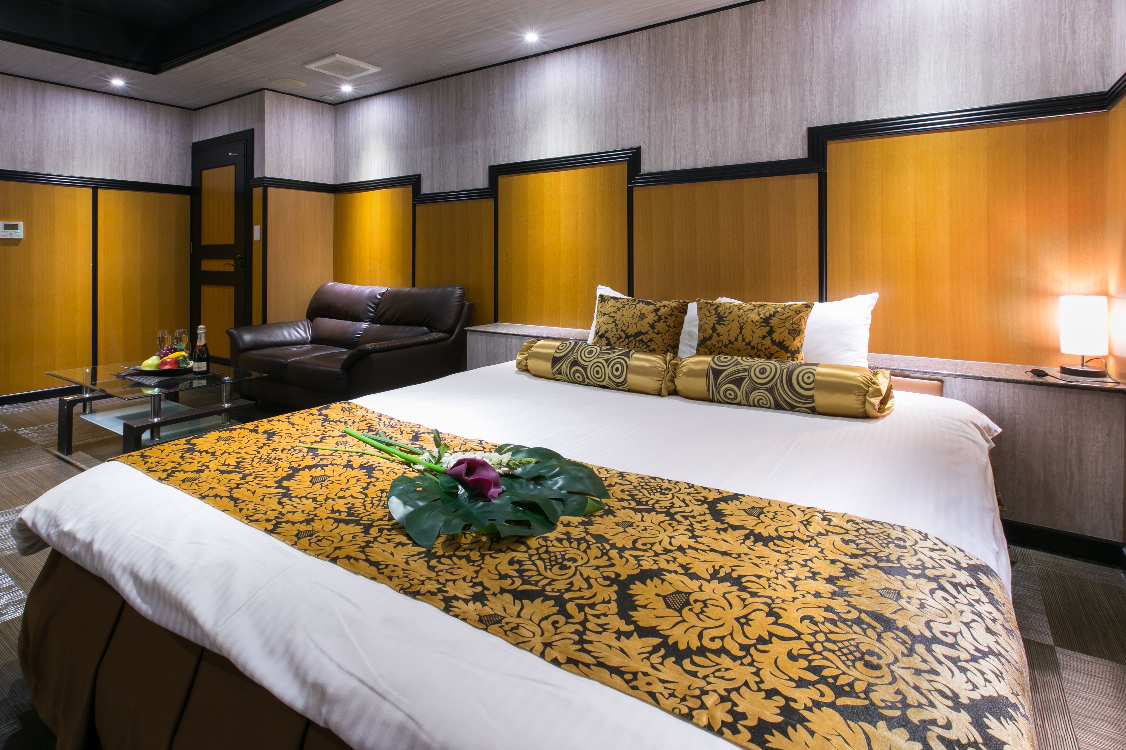 A hotel room with a large bed, a leather armchair, a bedside table with a lamp, and a small table with a fruit bowl and bottle.